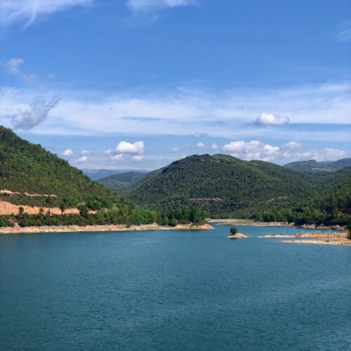 View of the Rialb reservoir in the background mountains with their leafy forests. Blue sky with some clouds that look like cotton wool.