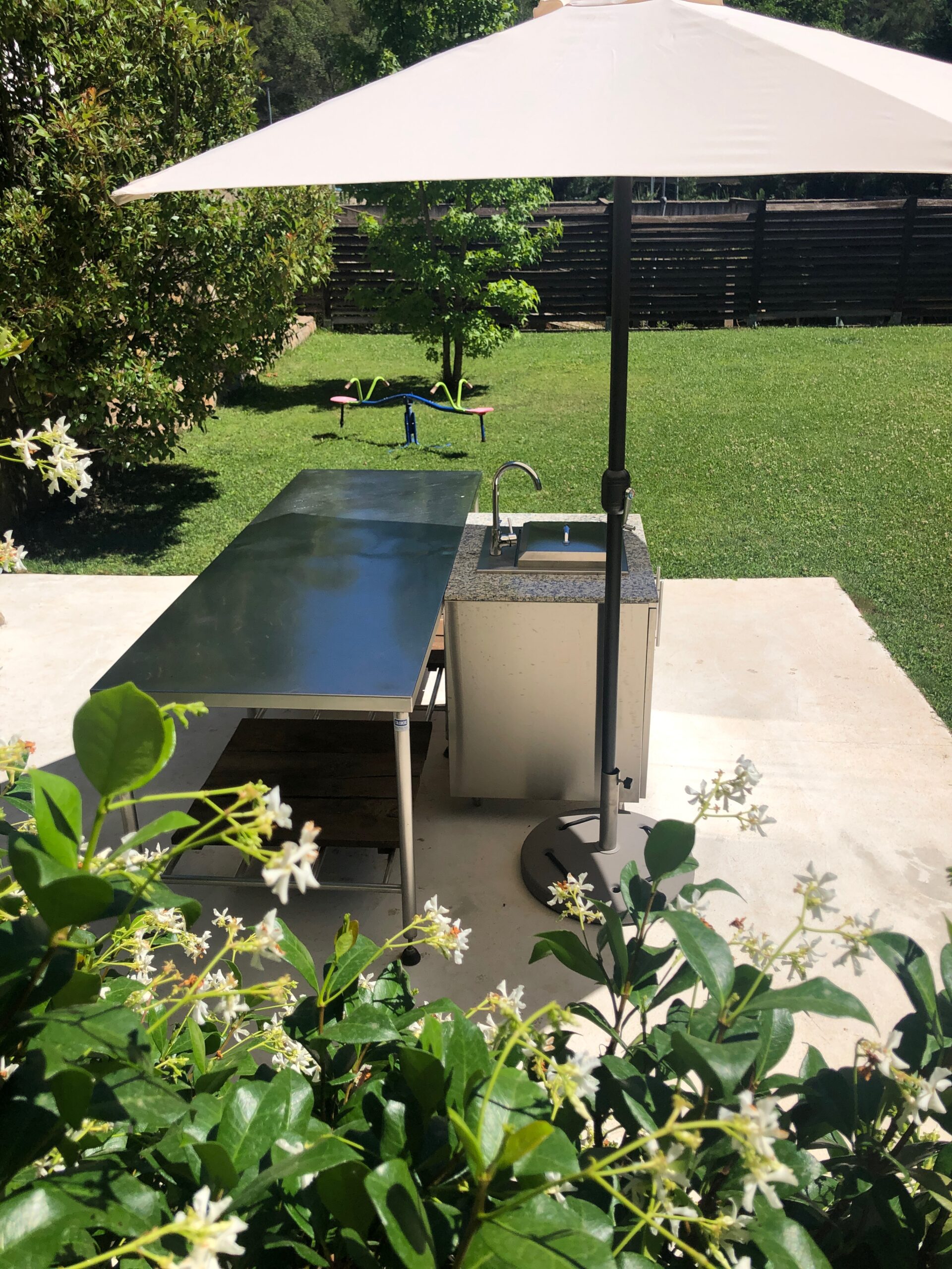 Detail of the outdoor barbecue with large stainless steel table in the center and furniture with kitchen sink and large umbrella. In the background the garden, you can also see a jasmine vine with white flower.