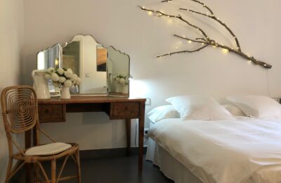 Habitación donde se puede ver una parte de la cama con ropa blanca y cojines mullidos al lado un tocador con espejo de mesa de tres caras a la pared original lámpara con una rama de árbol seca.