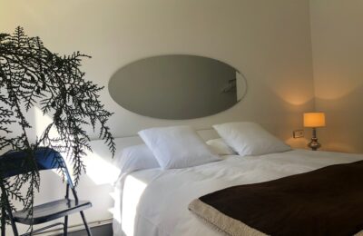 Habitación cama doble ropa de cama blanca con manta oscura en los pies silla de metal espejo ovalado en el cabezal, adorno con hojas de abeto natural