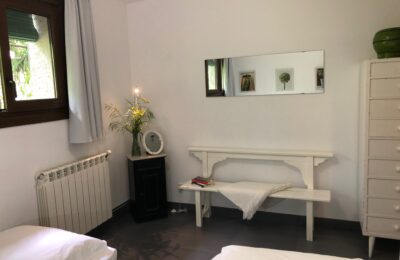 Detalle habitación con dos camas individuales al fondo banco de madera blanco encima en la pared espejo rectangular, al lado mesita de noche negra con espejo obalado pequeño y un jarrón con un ramo de flores blancas