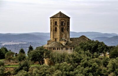 Preciosa vista de Sant Pere de Ponts,rodeada de vegetación de la zona, pinos robles y encinas , al fondo las montañas. De la colegiata se puede ver la torre principal y dos de sus tres ábsides