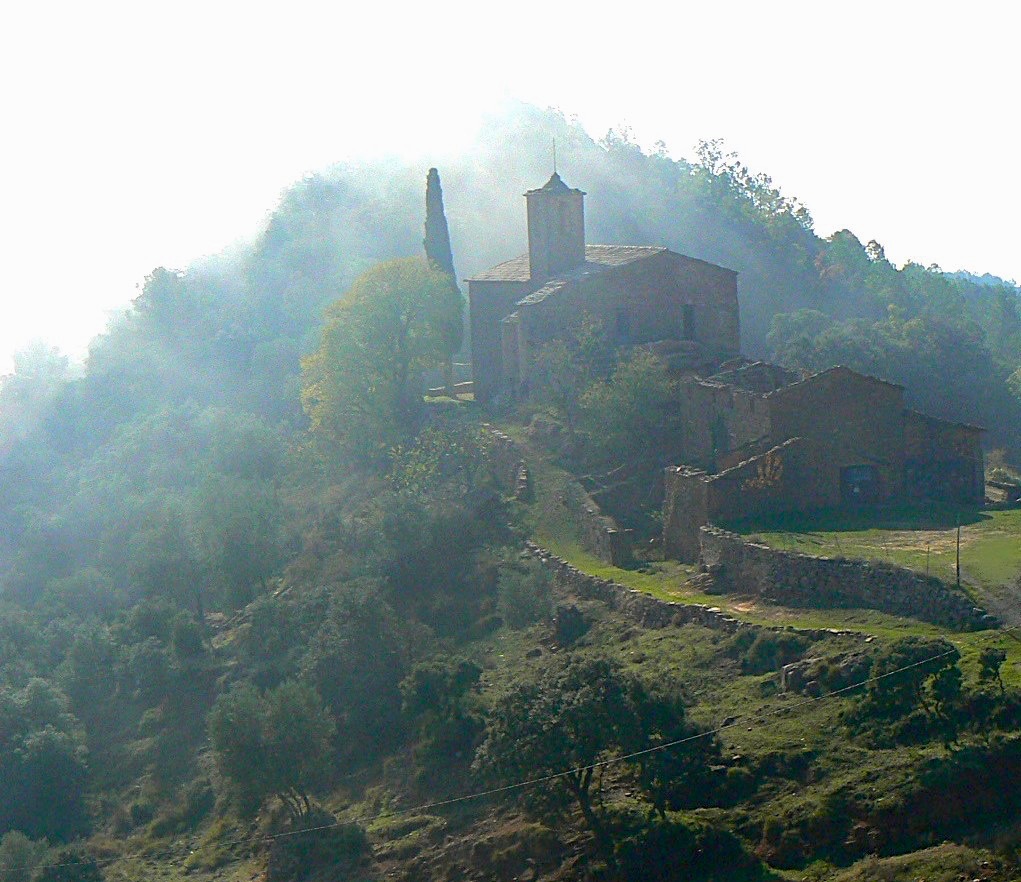 Vista de la iglesia de Pallerols ubicada encima de un monticulo con un ciprés al lado derecho y la niebla que parece que se mueve