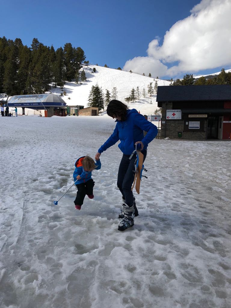 Llano del final de una pista de esquí, niño jugando con adulto. Un cielo azul brillante con unas motas de nubes blancas que parecen algodones.Al fondo se aprecia la pista de esquí.