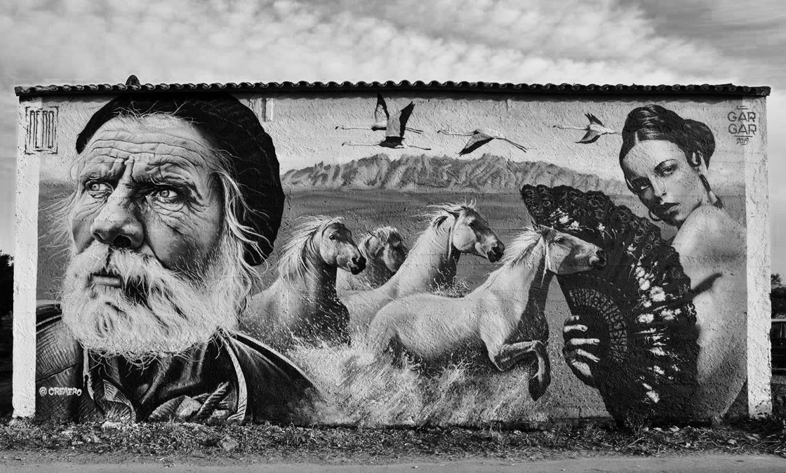Photo of a graffiti in the town of Penelles. A tanned face of an adult man, in the centre three white horses galloping through the water, a woman characterised as a Sevillian woman with a fan covering her naked torso and finally three storks in flight. Painted in black and white and grey.