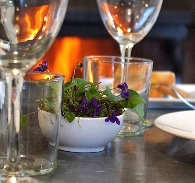 Copas de vino encima la mesa, entre ellas un ramillete pequeño de violetas, al fondo chimenea encendida. Todo en su sitio para empezar a degustar la cena junto una buena copa de vino.